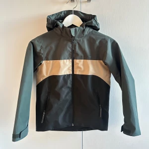 Grön och beige vindjacka med huva barn - Snygg grön och beige vindjacka med huva och dragkedja framtill. Jackan har långa ärmar och praktiska fickor med dragkedjor. Perfekt för blåsiga dagar.