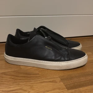 Svarta sneakers från Axel Arigato - Snygga svarta sneakers i skinn från Axel Arigato med dragkedja på ovansidan och vit sula. Perfekta för en stilren look. Inget og finns bara skorna 