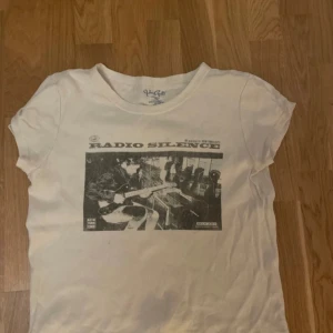 Vit t-shirt med tryck från Radio Silence - Säljer en vit t-shirt från Radio Silence med ett coolt svartvitt tryck på framsidan. T-shirten har korta ärmar och en rund halsringning. Perfekt för en avslappnad stil.