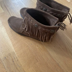 Bruna mocka boots med fransar - Snygga bruna boots i mocka med fransdetaljer och flätad kant. Skorna har dekorativa knappar och en dragkedja på sidan för enkel på- och avtagning. Perfekta för en bohemisk stil.
