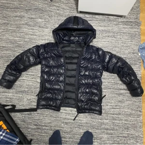 Svart dunjacka från Peak Performance - Säljer en svart dunjacka från Peak Performance. Jackan har en huva och dragkedja framtill. Perfekt för kyligare väder med sin pufferdesign som håller värmen. Märkeslogga synlig på ärmen. Storlek 160 Men en lagning lite ovanför fickan men syns inte för armen är över.