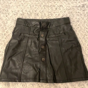 Svart kjol från Lindex - Snygg svart kjol i skinnimitation från Lindex med knappar framtill. Perfekt för en cool look.