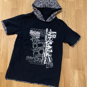 Svart kortärmad hoodie från Airwalk y2k vtg - Säljer en svart kortärmad hoodie från Airwalk med tryck i vitt och grått. Hoodien har en mönstrad huva och ärmslut med textdetaljer. Perfekt för en avslappnad stil.