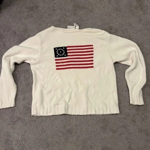 Vit stickad tröja med amerikansk flagga - Säljer en vit stickad tröja med en amerikansk flagga på framsidan. Tröjan har långa ärmar och ribbstickade muddar vid ärmslut och nederkant. Perfekt för en avslappnad stil.
