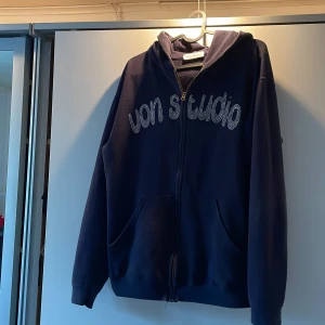 Mörkblå hoodie från Von studio - Snygg mörkblå hoodie från Vain Studio med dragkedja och glittrig text på framsidan. Baksidan har ett coolt motiv i samma stil. Perfekt för en avslappnad stil. Passar dig som bär storlek M-L. Denna tröja från von studio är limited edition och säljs därför inte nånstans längre. Köpt för 1500kr. Denna modell är alltså extremt sällsynt.