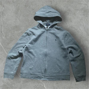 Grå hoodie från Ralph Lauren - Säljer en stilren grå hoodie från Ralph Lauren med dragkedja och klassisk logga på bröstet. Tröjan har en bekväm passform och är perfekt för kyligare dagar. Den har en praktisk huva och fickor framtill.