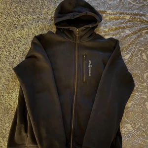 Svart fleecehoodie från Sail Racing - Säljer en svart zip hoodie från Sail Racing med huva samt flera fickor. Koftan har en bröstficka med dragkedja, samt två vanliga fickor och är perfekt för kyliga dagar. Den är långärmad och har en stilren design. Säljer då den inte kommer till användning längre. Nypris 1299kr😊