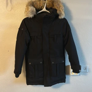 Svart parkajacka med päls från Nobis - Säljer en stilren svart jacka från Nobis med avtagbar päls på huvan. Nypris på jackan ligger på 10 000kr. Jackan har flera praktiska fickor med knappar och en diskret logga på ärmen. Perfekt för kalla dagar. Storlek L barn. Priset kan diskuteras!