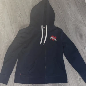 Mörkblå hoodie från Tommy Hilfiger - Säljer en snygg mörkblå hoodie från Tommy Hilfiger med dragkedja och huva. Den har ett rött hjärta med texten 'Tommy' på bröstet och vita snören. Perfekt för en avslappnad stil.