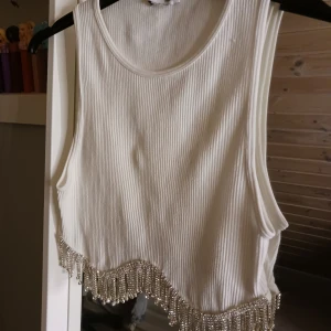 Vit ribbad linnetopp med fransar - Snygg vit ribbad linnetopp med glittriga fransar längs nederkanten. Perfekt för en festlig look. Toppen är ärmlös och har en rund halsringning. Köpt på plik men aldrig använd av mig😊
