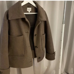 Brun kappa från Kappahl - Snygg brun kappa från Kappahl med stora fickor och knappar framtill. Jackan har en klassisk krage och långa ärmar med knappdetaljer vid ärmsluten. Perfekt för höst och vår!