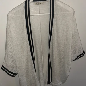 Vit kimono med svarta detaljer - Snygg och luftig vit kofta/kimono från Bershka med svarta kanter. Perfekt för lager-på-lager och ger en stilren look. Koftan har korta ärmar och en öppen design.