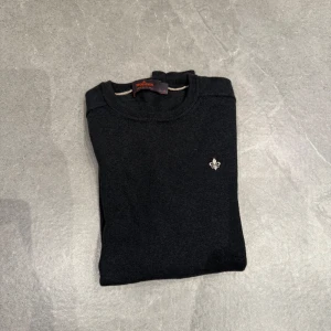 Svart merinoulltröja från Morris - Snygg svart tröja från Morris i 100% merinoull. Den har en liten broderad detalj på bröstet och ribbade kanter vid ärmslut och nederkant. Perfekt för kyligare dagar.