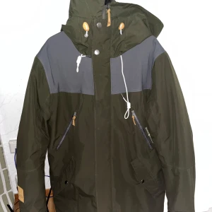 Ahkka outdoor jacka - Snygg grön parkajacka med grå detaljer och praktisk huva. Jackan har dragkedja och knappar framtill samt flera fickor med dragkedjor. Perfekt för kyligare väder. Storlek M knappt använd 