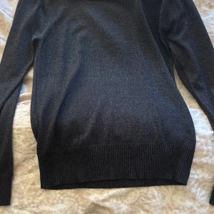 Svart glittrig tröja - Snygg svart tröja med glittrande effekt. Perfekt för att lägga till lite glamour i vardagen. Tröjan har långa ärmar och en ribbad nederkant som ger en bekväm passform.
