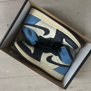 Nike Air Jordans i blått och vitt - Snygga Nike Air Jordans i en stilren kombination av blått, vitt och svart. Skorna har en mid-hög modell med snörning och den klassiska Nike-loggan på sidan. Modellen ”Obsidians” och i strl 38.5, i bra skick och använda fåtal gånger. 