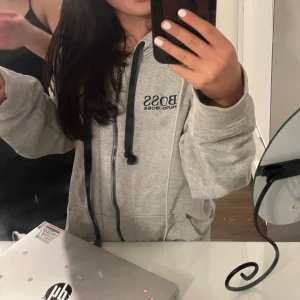 Grå hoodie från Hugo Boss - Säljer en stilren grå hoodie från Hugo Boss med dragkedja och justerbar huva. Tröjan har ett broderat emblem i rött, vitt och blått på sidan samt mörkblå snören. Det är en mans zip hoodie men funkar för båda könen❤️