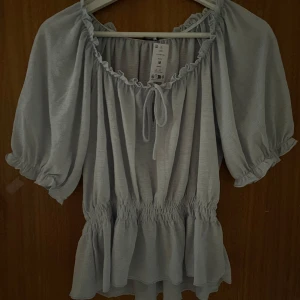 Ljusblå puffärm topp - Säljer en söt ljusblå topp med puffärmar och volangdetaljer. Toppen har en knytning framtill och elastisk midja för en bekväm passform. Aldrig använd - lappen är kvar.