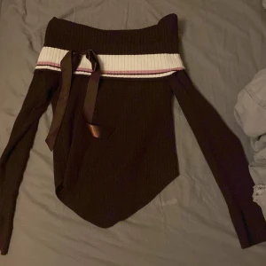 Brun offshoulder tröja med rosett - Snygg brun offshoulder tröja med ribbad design och dekorativ rosett framtill. Tröjan har en vit och rosa randdetalj vid axlarna som ger en fin kontrast. Perfekt för en stilren look.