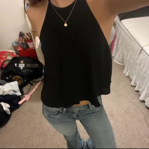 Svart ärmlös topp - Snygg svart ärmlös topp med halterneck-design. Perfekt för en avslappnad stil, lätt att matcha med jeans eller kjol. Toppen har en lös passform och är tillverkad i ett mjukt material.