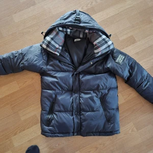 Svart dunjacka från Burberry - Snygg svart dunjacka från Burberry med klassiskt rutigt foder i huvan. Jackan har dragkedja och knappar framtill samt en stilren patch på ärmen. Perfekt för kyliga dagar. Jackan är använda en månad så perfekt skick 