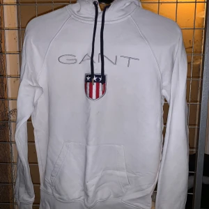 Vit hoodie från GANT - Säljer en stilren vit hoodie från GANT med broderad logga och ett emblem på bröstet. Hoodien har en klassisk känguruficka och justerbar dragsko i huvan. Perfekt för en avslappnad stil. 