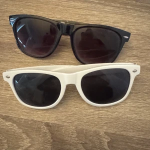 Svarta och vita solglasögon - Två par solglasögon i klassisk stil. Ett par är helsvarta med mörka linser och det andra paret är vita med mörka linser. Perfekta för soliga dagar och ger en stilren look.