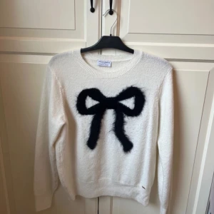 Vit tröja med svart rosett från Holly & Whyte - Säljer en vit tröja från Holly & Whyte med en stor svart rosett på framsidan. Tröjan har långa ärmar och är tillverkad i ett mjukt material.