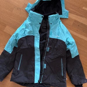 Turkos och svart vinterjacka från Everest - Säljer en snygg turkos och svart vinterjacka från Everest. Jackan har en praktisk dragkedja och knappar framtill, samt en justerbar huva. Perfekt för kalla dagar med sina långa ärmar och flera fickor med dragkedjor.