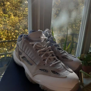 Air Jordan 11 Retro Low IE - Snygga Air Jordan 11 Retro Low IE i en stilren kombination av ljusbrun och grå. Skorna har en unik design med meshdetaljer och snörning. Perfekta för dig som vill ha en sportig och trendig look.