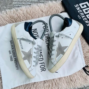 Golden Goose sneakers med stjärndetalj - Snygga vita sneakers från Golden Goose med grå stjärndetalj på sidan och mörkblå häl. Skorna har en vintage-look med snörning och en bekväm gummisula. Perfekta för en avslappnad stil.