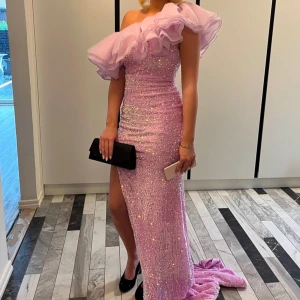 Rosa/lila glittrig balklänning - Säljer en fantastisk rosa/lila balklänning med glittrande paljetter och en dramatisk volangdetalj vid axlarna. Klänningen har en slits framtill och en elegant offshoulder-design. Perfekt för balen eller en glamourös kväll! Den är endast använd fåtal gånger! Bra skick🤩Dregkedjan i ryggen är rätt trög så man måste kämpa lite att få upp den men tillslut går det!