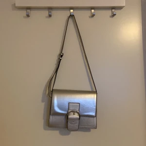 Silver axelväska från Orsay - Snygg silverfärgad axelväska från Orsay med justerbar rem och stilren design. Väskan har en stor dekorativ spänne framtill och ett rymligt inre fack. Aldrig använd! 