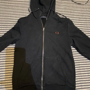 Svart hoodie från Fred Perry - Hej grabbar. Säljer en stilren svart hoodie från Fred Perry med dragkedja och huva. Tröjan har långa ärmar och en liten logotyp på bröstet. Perfekt för en avslappnad look. Ett litet hål vid fikan.7/10 kvalitet.      Priset är inte hugget i sten