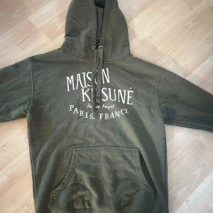 grön hoodie från Maison Kitsuné - Säljer en snygg och väldigt unik grön hoodie från Maison Kitsuné med tryck på framsidan. Den har en klassisk känguruficka och justerbar huva med snören. Perfekt för en avslappnad stil. Nypris ca 3000 mitt pris 899. Använd 4 ggr, pris kan diskuteras vid snabb affär