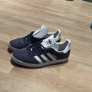 Adidas Gazelle i mörkblå mocka - Säljer ett par klassiska Adidas Gazelle i mörkblå mocka med vita detaljer och snörning. De följer även med ett par vita snören.hör av er om ni har några frågor 