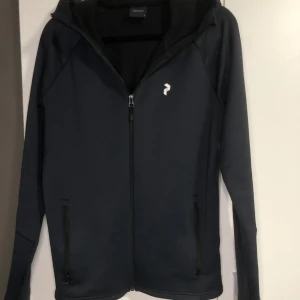Svart huvjacka från Peak Performance - Snygg svart huvjacka från Peak Performance med dragkedja framtill och en liten logga på bröstet. Jackan har långa ärmar och en bekväm huva. Perfekt för en sportig look. Finns ett stygn som gått upp på ena ärmen men annars i mycket bra skick. Köpt för 1500kr på stadium.