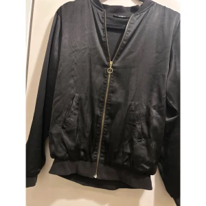 Svart satin bomberjacka med dragkedja, strl xs  - Snygg svart bomberjacka med dragkedja framtill. Tunn i materialet. Jackan har ribbade muddar vid ärmslut och nederkant. Strl XS!