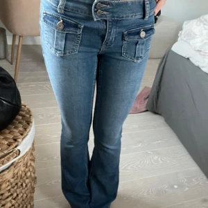 Blå jeans med fickdetaljer - Snygga blå jeans med unika fickdetaljer och knappar framtill. De har en lågmidjad passform och är bootcut! De är aldrig använda och nypriset är 2500kr
