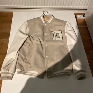 Beige och vit bomberjacka med broderi - Säljer en snygg beige och vit bomberjacka med broderad text 'Athletics Division League Tournament 1993' och ett stort 'D' för Detroit. Jackan har ribbade muddar och knappar framtill. Perfekt för en sportig look!