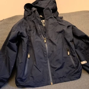 Mörkblå jacka från Kaxs - Säljer en mörkblå jacka från Kaxs med huva och dragkedja. Jackan har praktiska fickor med dragkedjor och reflexdetaljer på ärmarna. Perfekt för utomhusaktiviteter.