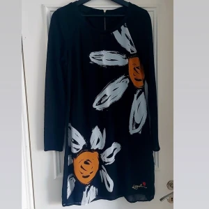 Svart klänning med blommönster från Desigual - Snygg svart klänning från Desigual med stora blommor i vitt och orange. Klänningen har långa ärmar och en avslappnad passform. Perfekt för att sticka ut med ett unikt mönster. 🌼
