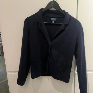 Mörkblå kavaj från Polo Ralph Lauren - Snygg mörkblå kavaj från Polo Ralph Lauren med knappar framtill och två fickor. Kavajen har långa ärmar och en klassisk krage. Kavajen har inte används så mycket, bara några gånger, och den är i bra skick 