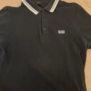 Svart pikétröja från Hugo Boss - Snygg svart pikétröja från Hugo Boss med vit randig krage och knappar framtill. Perfekt för en stilren look. Regular fit för en bekväm passform. Strl M men kan passa S, 350 kr är mitt sista pris, helt äkta och inga defekter. Kan mötas i Karlstad/Linköping, kan också fraktas (Köparen står för frakt). 
