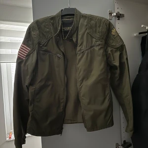 Grön jacka från Ralph Lauren - Säljer en grön jacka från Ralph Lauren med coola detaljer som dragkedjor och patchar på ärmarna. Jackan har en militärinspirerad look och är perfekt för höst och vår. Den har långa ärmar och en stilren design.