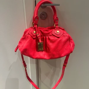 Marc Jacobs väska  - Snygg röd axelväska med gulddetaljer och justerbar axelrem. Väskan har en dragkedja upptill och ett mönstrat innerfoder med bokstäver. Perfekt för att ge en färgklick till din outfit!