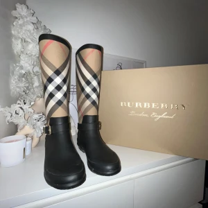 Rutig stövlar från Burberry - Snygga stövlar från Burberry med klassiskt rutig mönster i beige, svart och röd. Stövlarna har en dekorativ spänne och är perfekta för regniga dagar. De är både stiliga och praktiska. 