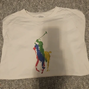 Vit t-shirt från Ralph Lauren med färgglatt tryck - Säljer en vit t-shirt från Ralph Lauren med ett stort, färgglatt tryck av en polospelare i blått, rött, grönt och gult. T-shirten har en klassisk passform och är perfekt för en avslappnad stil. Märket 'Polo' syns tydligt på etiketten. STRL S!!