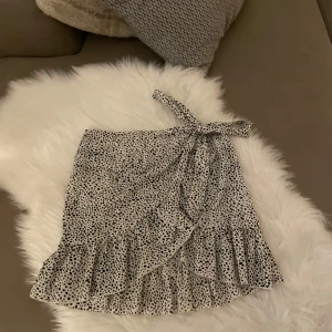 Prickig omlottkjol med volang - Snygg prickig omlottkjol i svart och vit med volangdetaljer. Kjolen har en knytning i midjan och är perfekt för en trendig look. Den är kort och passar bra till både vardag och festliga tillfällen.