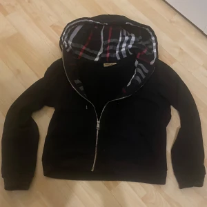 Svart hoodie från Burberry - Säljer en snygg svart hoodie från Burberry med klassiskt rutigt mönster i huvan. Jackan har dragkedja framtill och är tillverkad i bomull. Perfekt för en stilren look. Koftan är använd fåtal gånger.